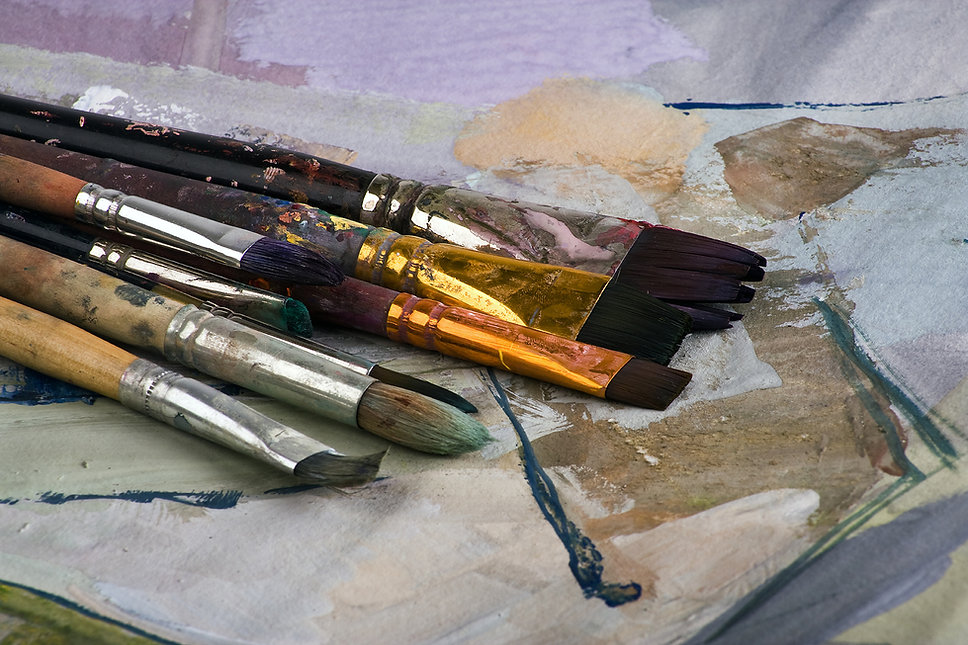 Close-up of used paintbrushes with paint-stained handles resting on a paint-splattered cloth, in muted earthy tones